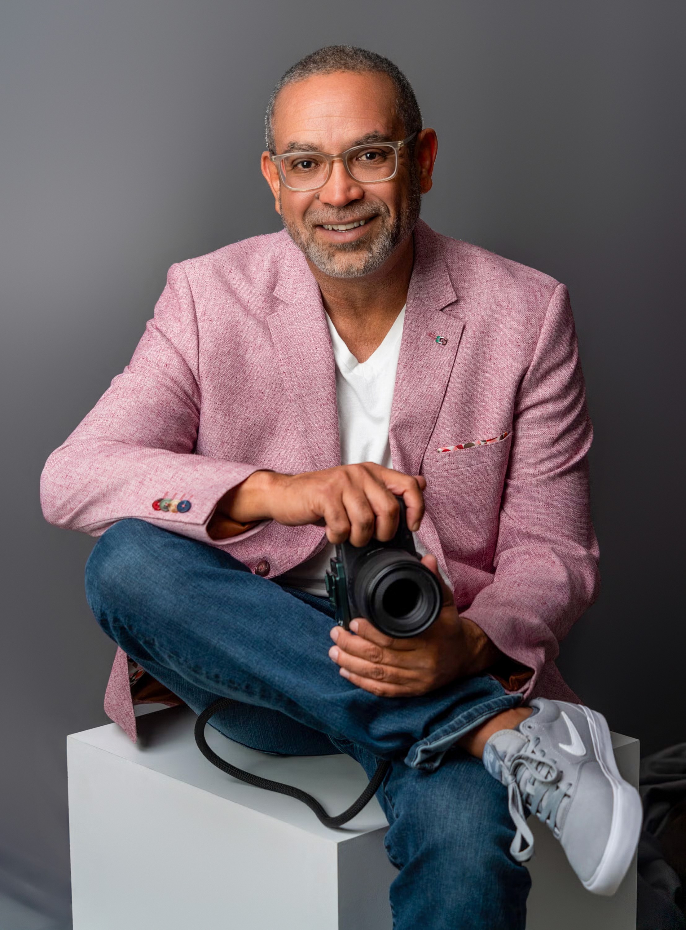 Portrait of Benjamin Hernandez seated with a camera in hand, smiling in a pink blazer, studio background.
