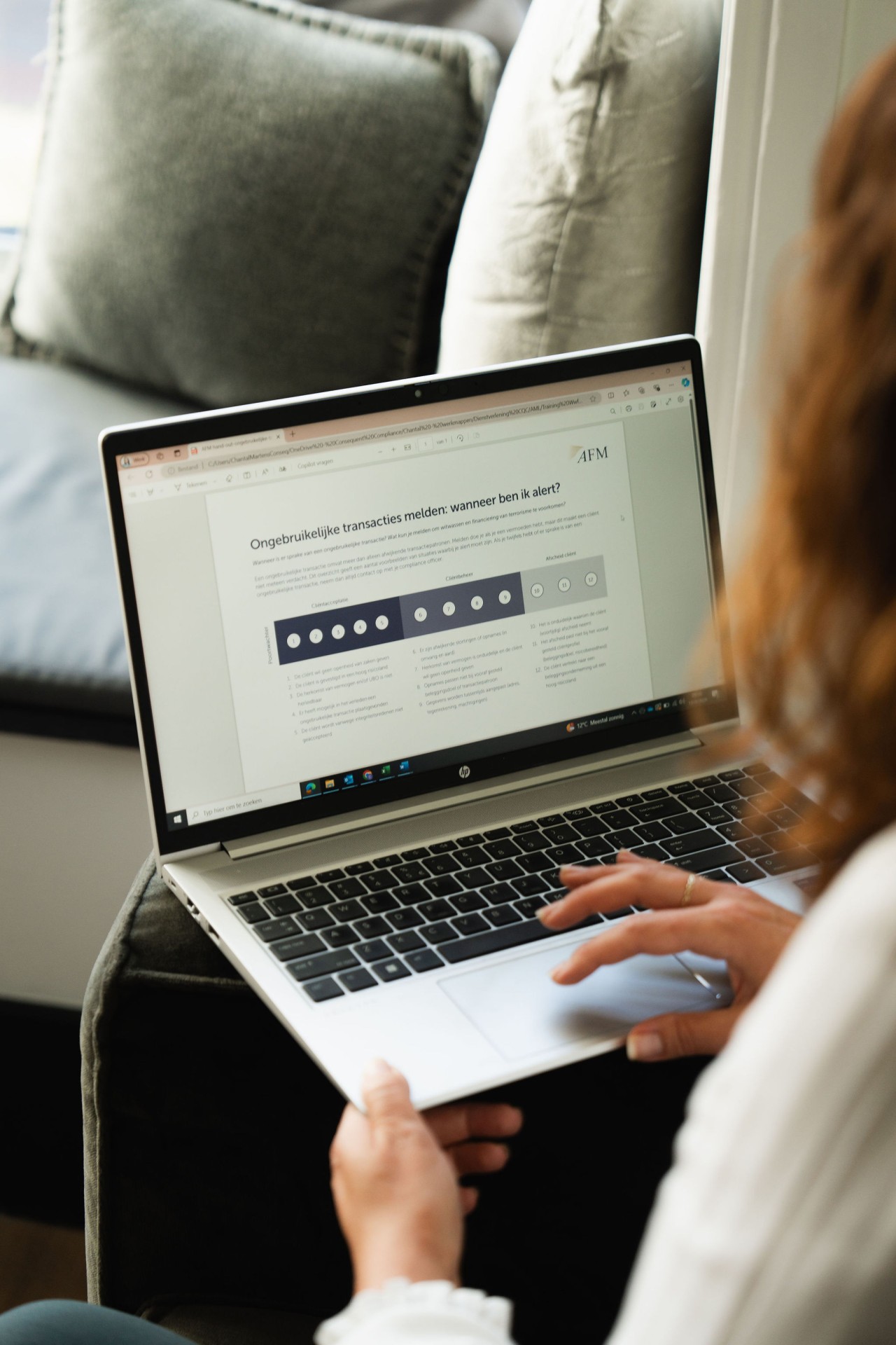 A man wearing glasses and a blazer working at a desk, writing in a notebook beside a computer monitor in a modern office.