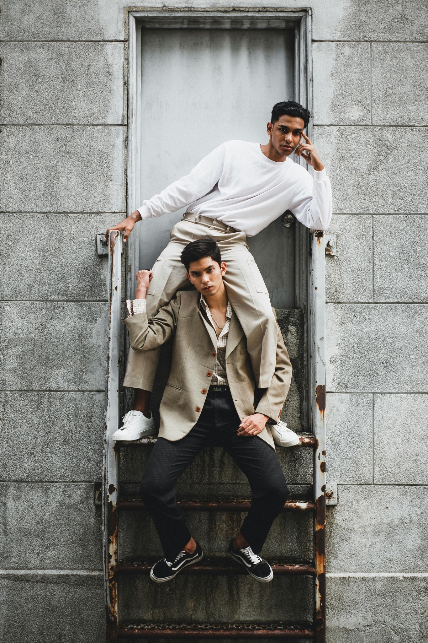 Two people are playfully posing, one perched on a ledge above the other, against a concrete wall.