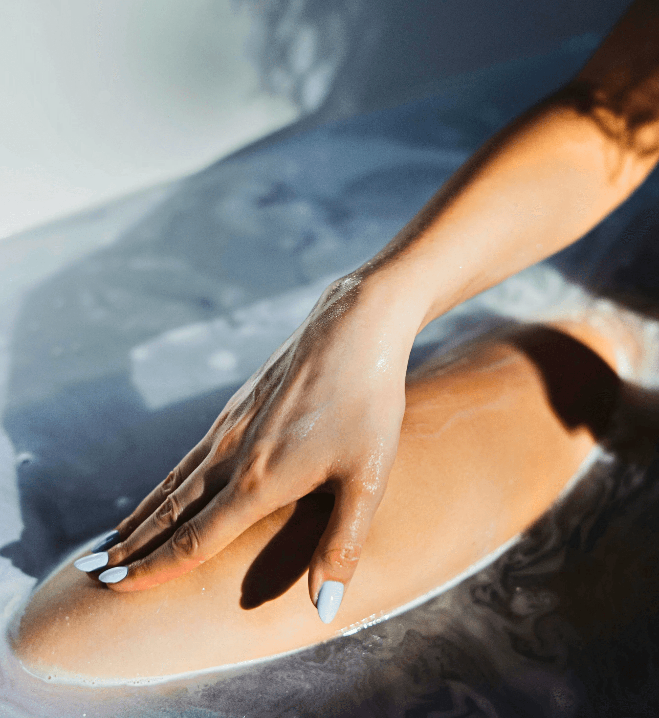 Close-up of a hand with white manicured nails gently resting on a leg submerged in a bath filled with water and bubbles, illuminated by soft natural light