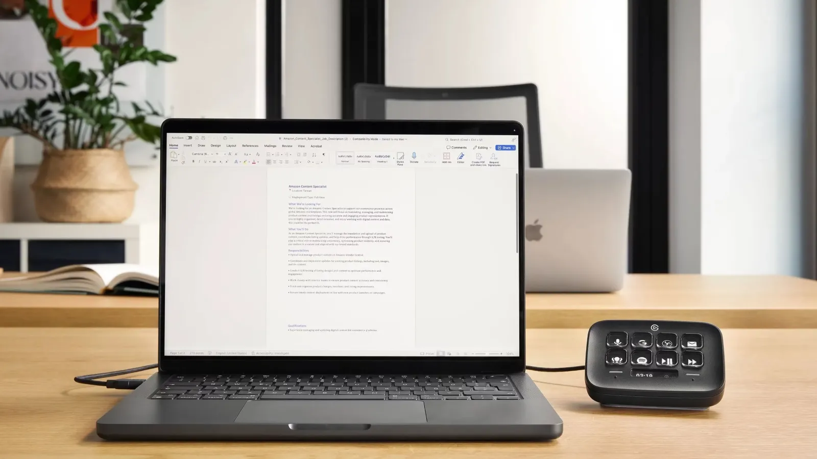 A sleek laptop displaying a document sits on a wooden desk beside an open book and a digital control panel, with a plant and another laptop in the blurred background, highlighting a modern workspace setting.