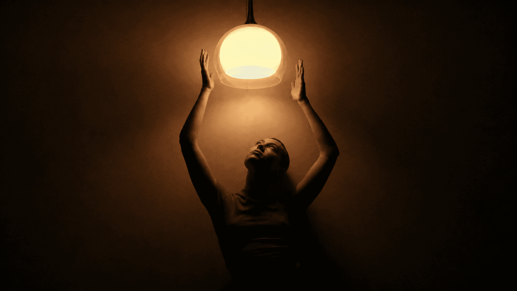 A woman in a dark room reaches up towards a glowing pendant lamp.