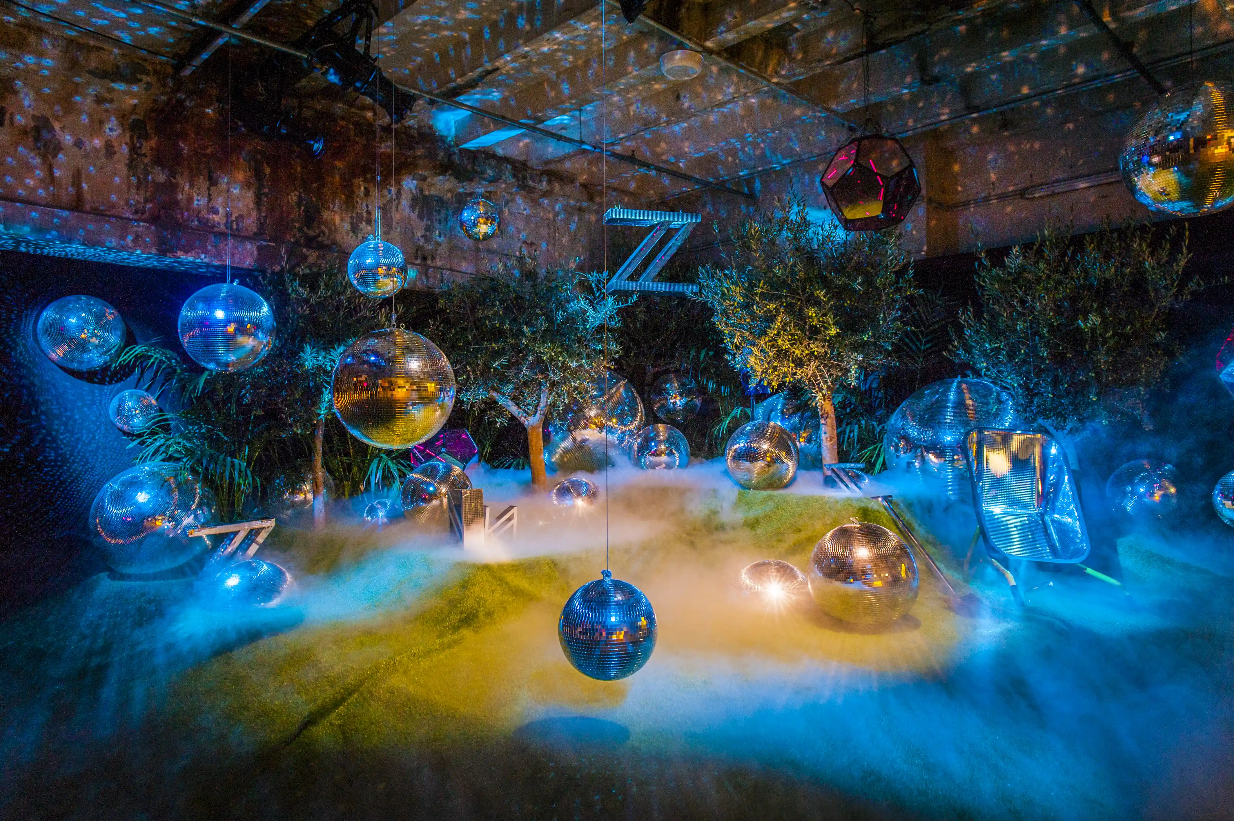 Dreamlike installation at BERLIN PHOTOWEEK with mirror spheres and geometric lanterns hanging over a misty mound with olive trees in blue light.