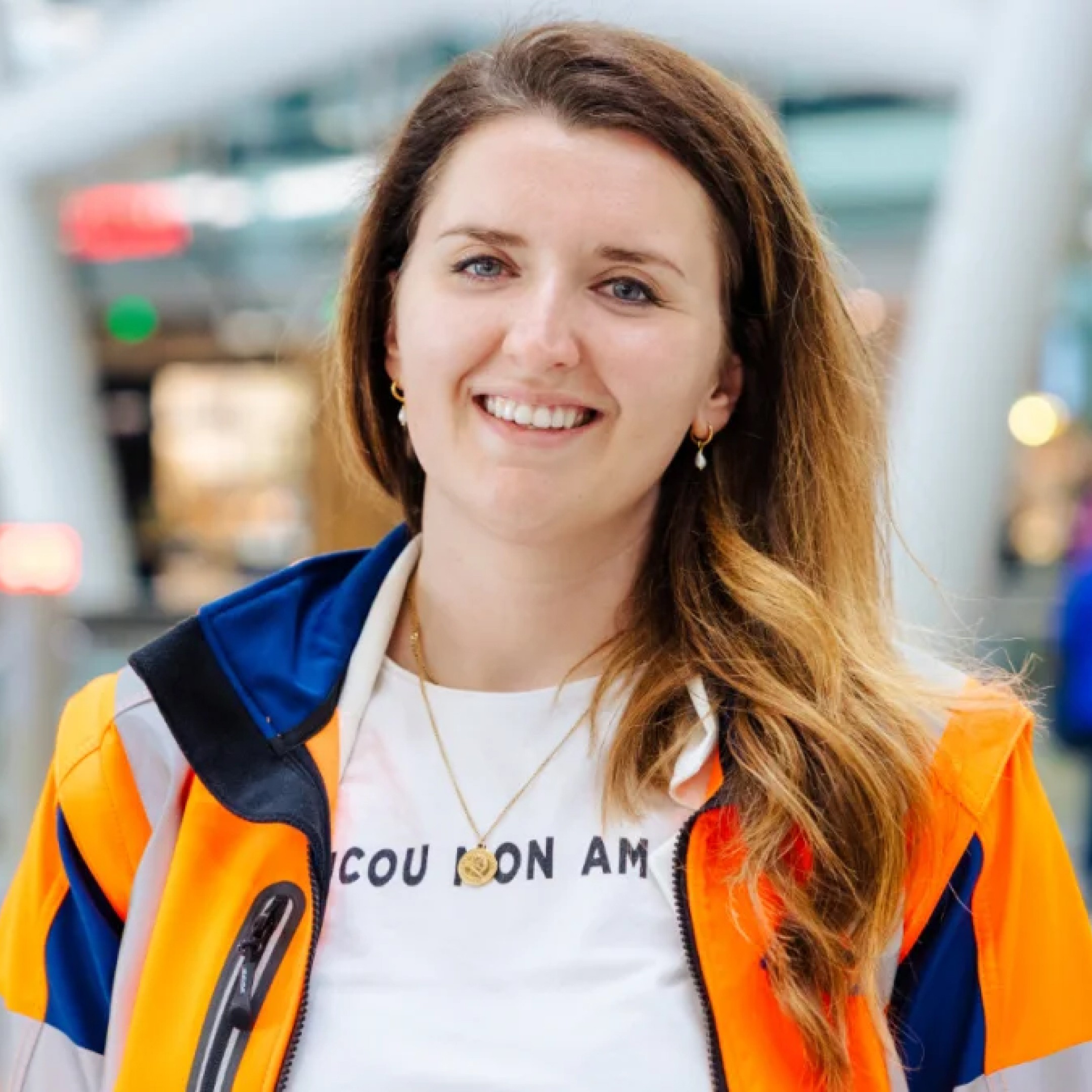 Corien Brouwer van de NS, lachend in een oranje veiligheidsjack en een wit T-shirt, poseert in een moderne omgeving.