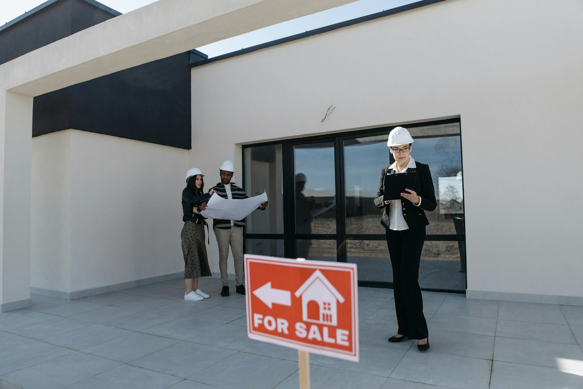 Three people in hard hats outside a modern white building with a red For Sale sign in the foreground