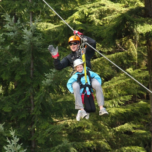 兩個人穿著頭盔和安全裝備，正在樹林中滑索。成人揮手，孩子微笑。