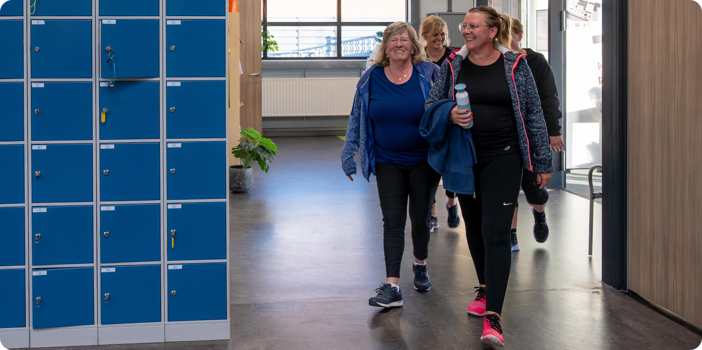 A group of smiling women casually walk through a gym corridor beside blue lockers, with one holding a water bottle and wearing workout attire, embodying a relaxed and social fitness atmosphere.