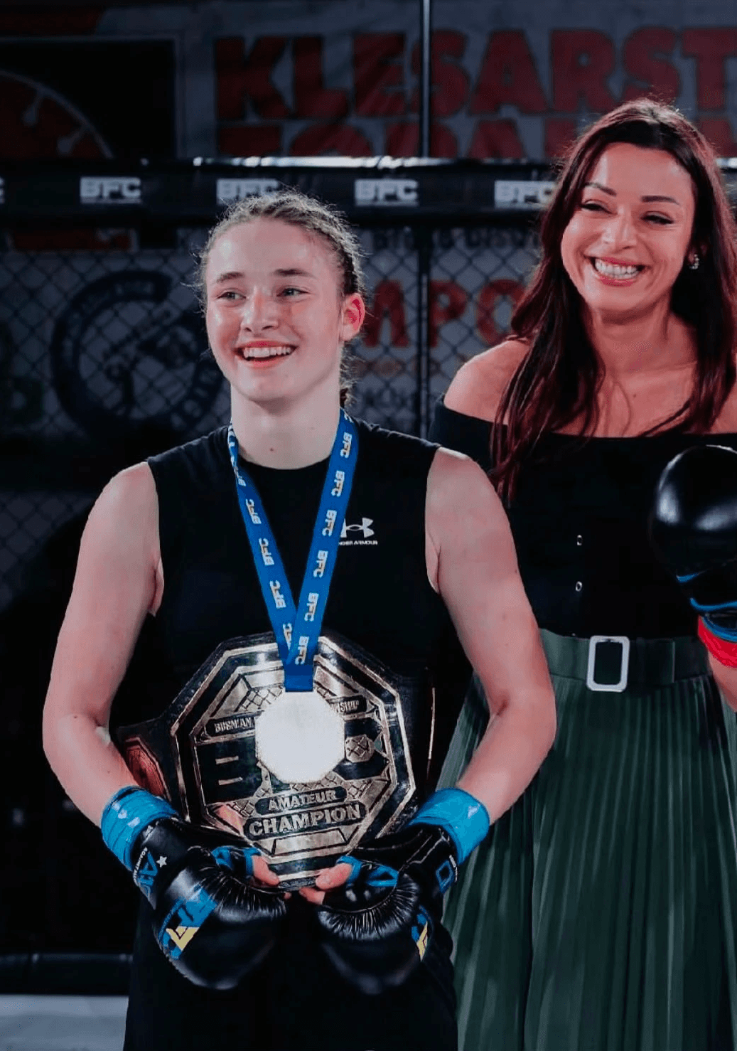 Mlada takmičarka pozira sa medaljom pored trenerice nakon nastupa na BFC takmičenju.
