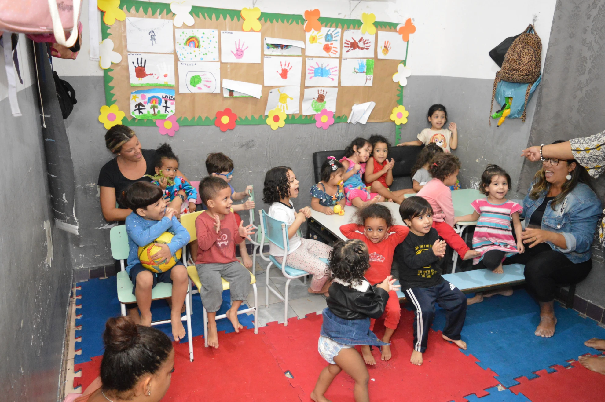 Grupo de crianças brincando e estudando em sala colorida, com cartazes educativos na parede.