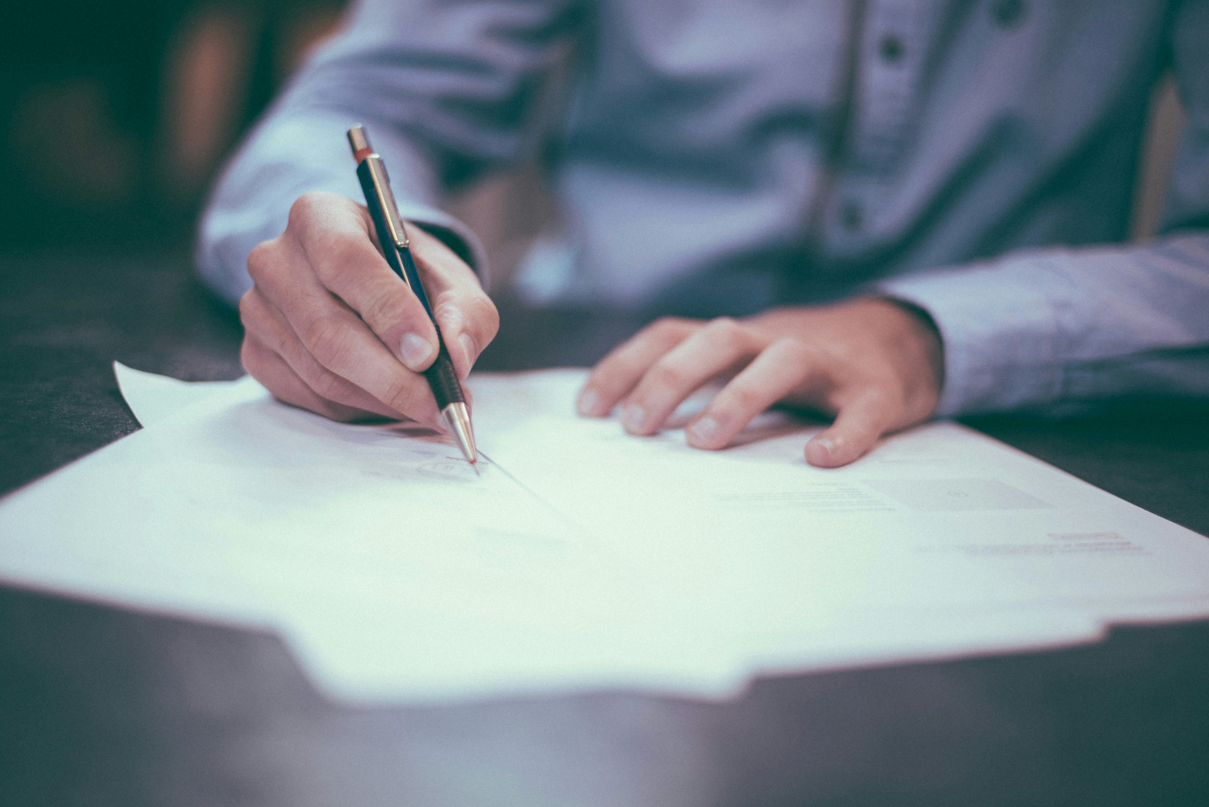 A person in a blue shirt is writing on several sheets of paper spread out on a wooden desk, with focus on the hand holding a pen, highlighting the concept of office work and documentation.