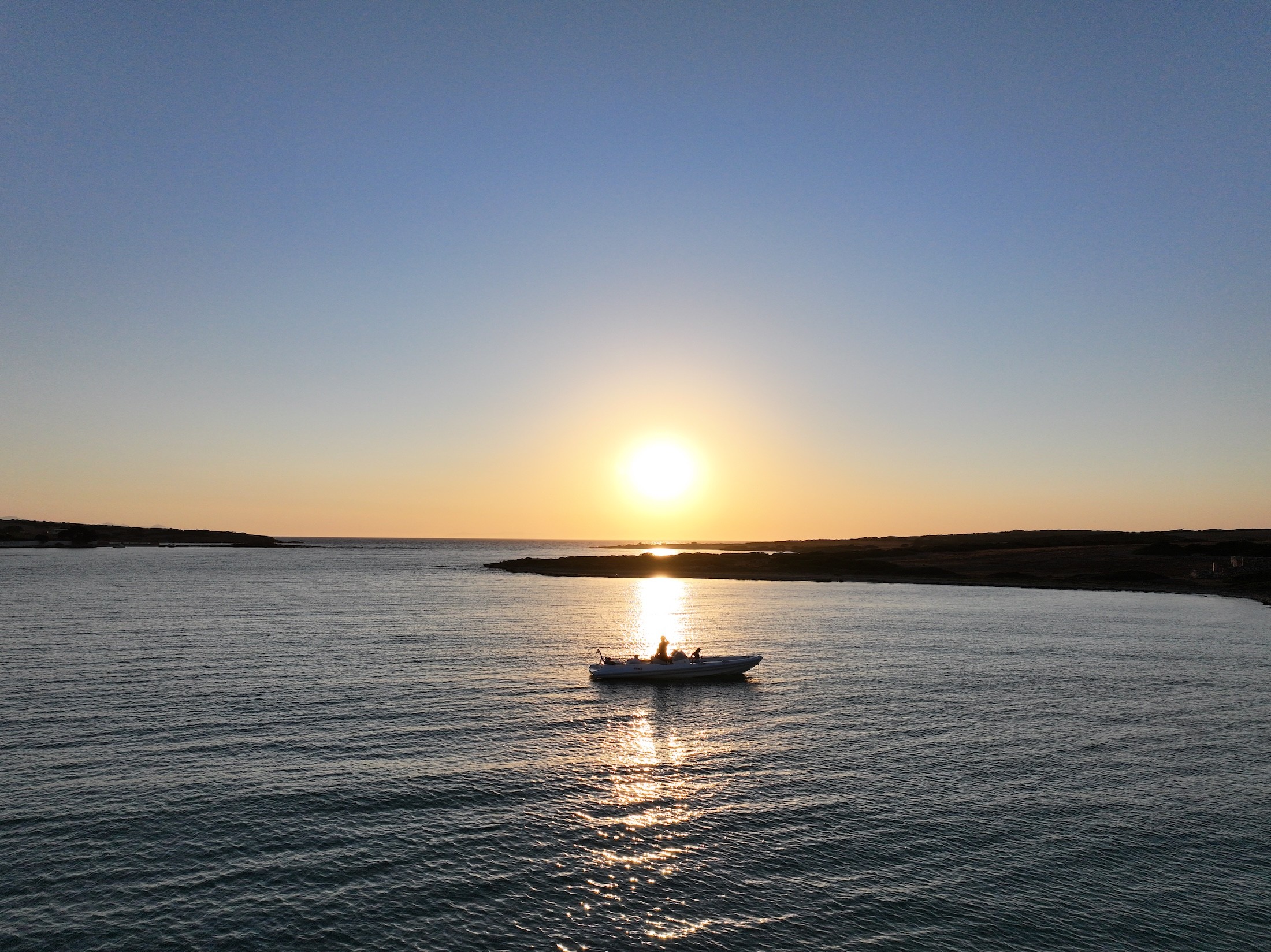 Small white motorboat silhouetted against golden sunset over calm Aegean waters with rocky coastline in background.