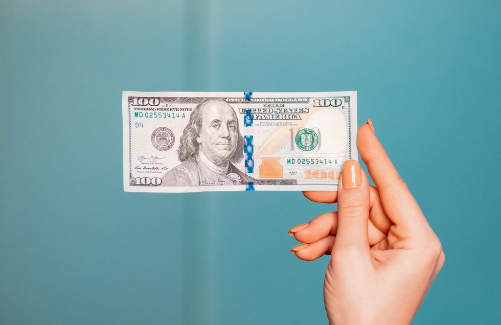 Hand holding a 100 dollar bill against a blue background.
