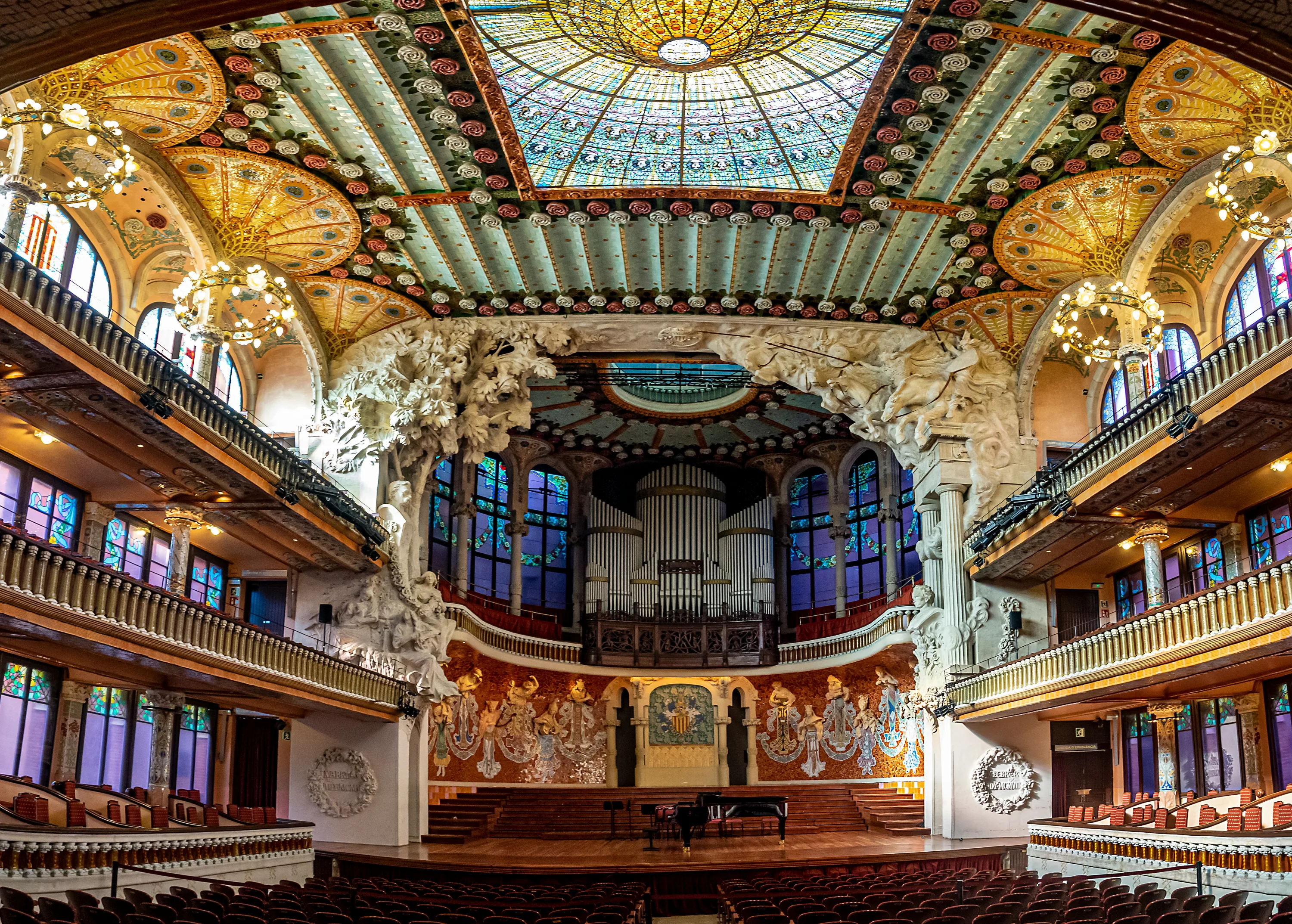 The ornate interior of the Palau de la Musica in Barcelona, featuring a bright stained glass skylight with a large pipe organ in the background raised above the stage.