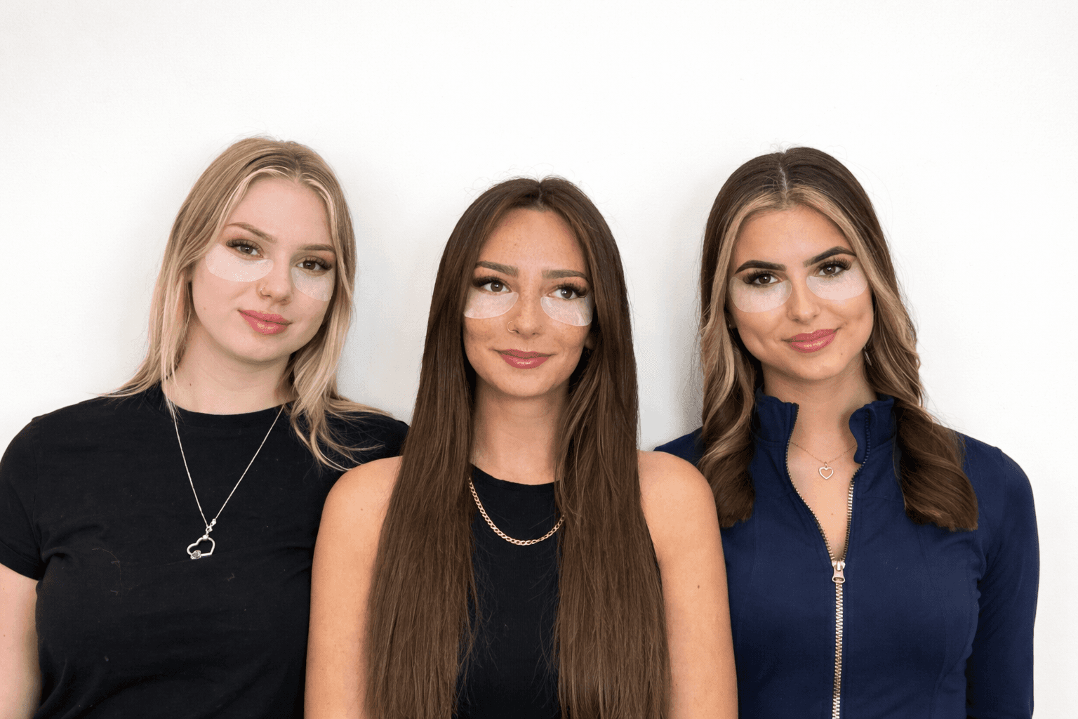 Three women stand together, smiling against a plain white background, wearing stylish outfits.