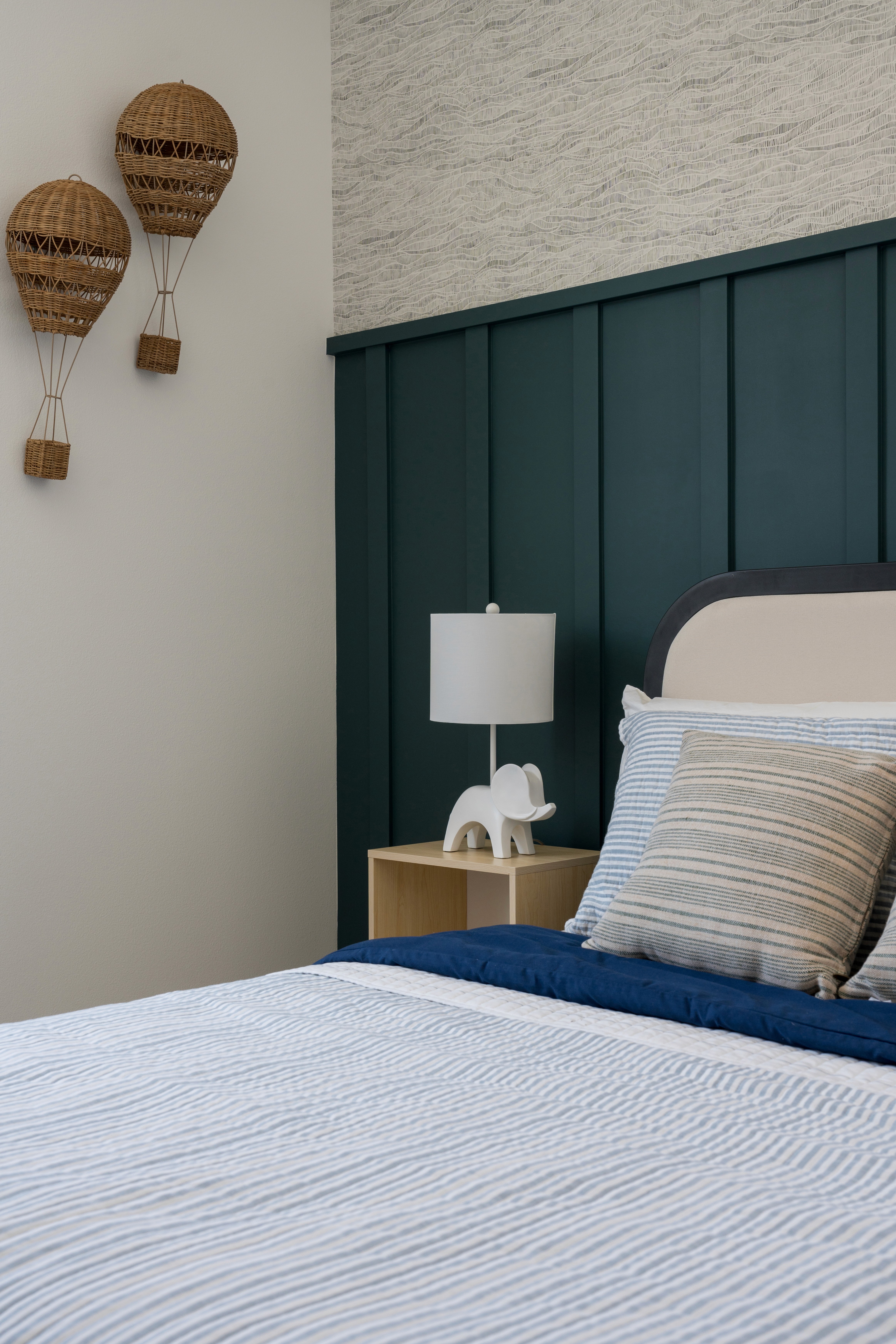 Boy's bedroom with light blue ribbed bedding, striped pillows, neutral headboard, deep blue wall paneling, and woven hot air balloon wall decor by Jubilee Interiors in Arcadia, California