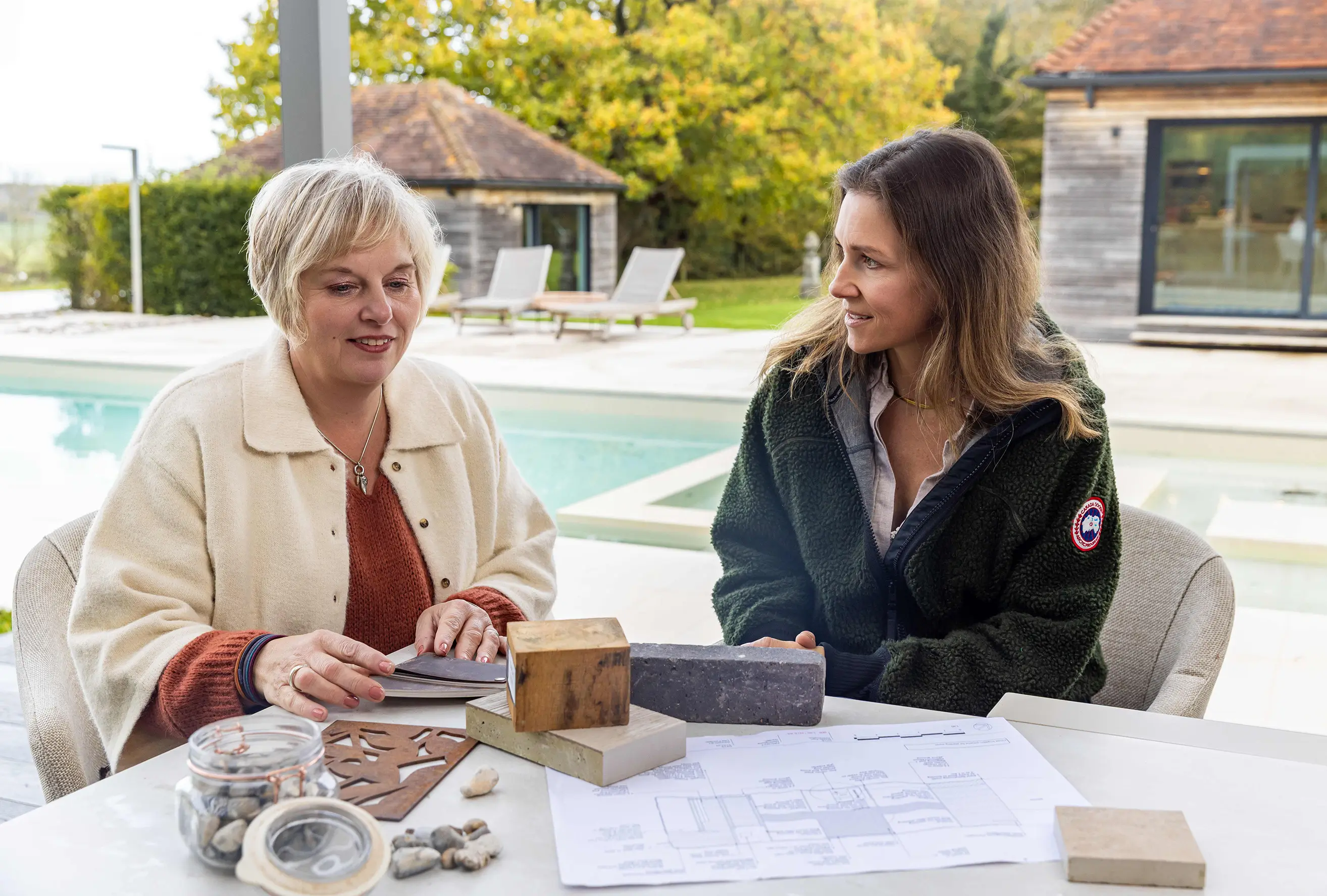 Two women sit at a table outdoors, engaged in conversation, with a pool and trees in the background.