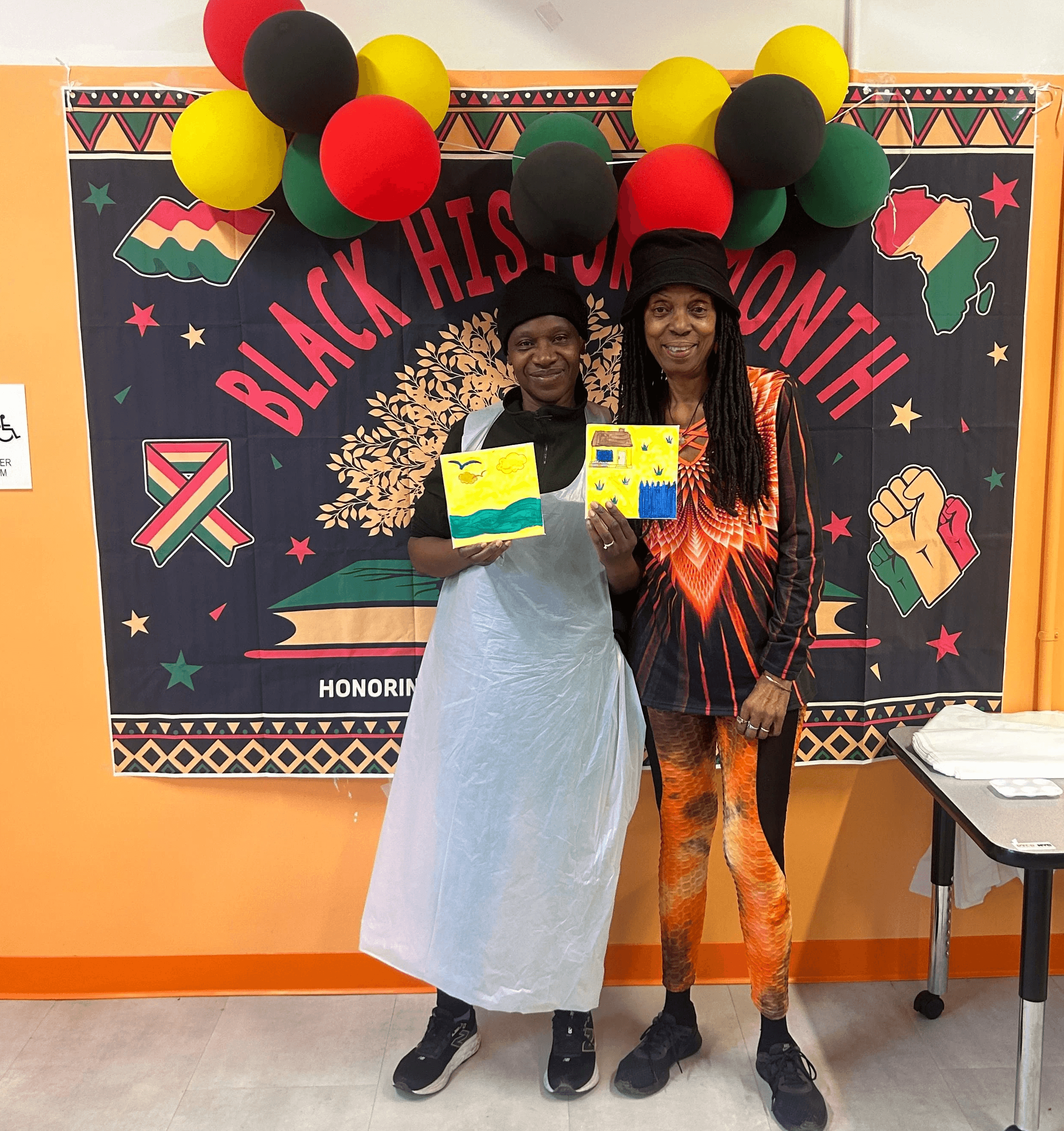 Two women stand in front of a Black History Month sign proudly displaying their artistic creations.