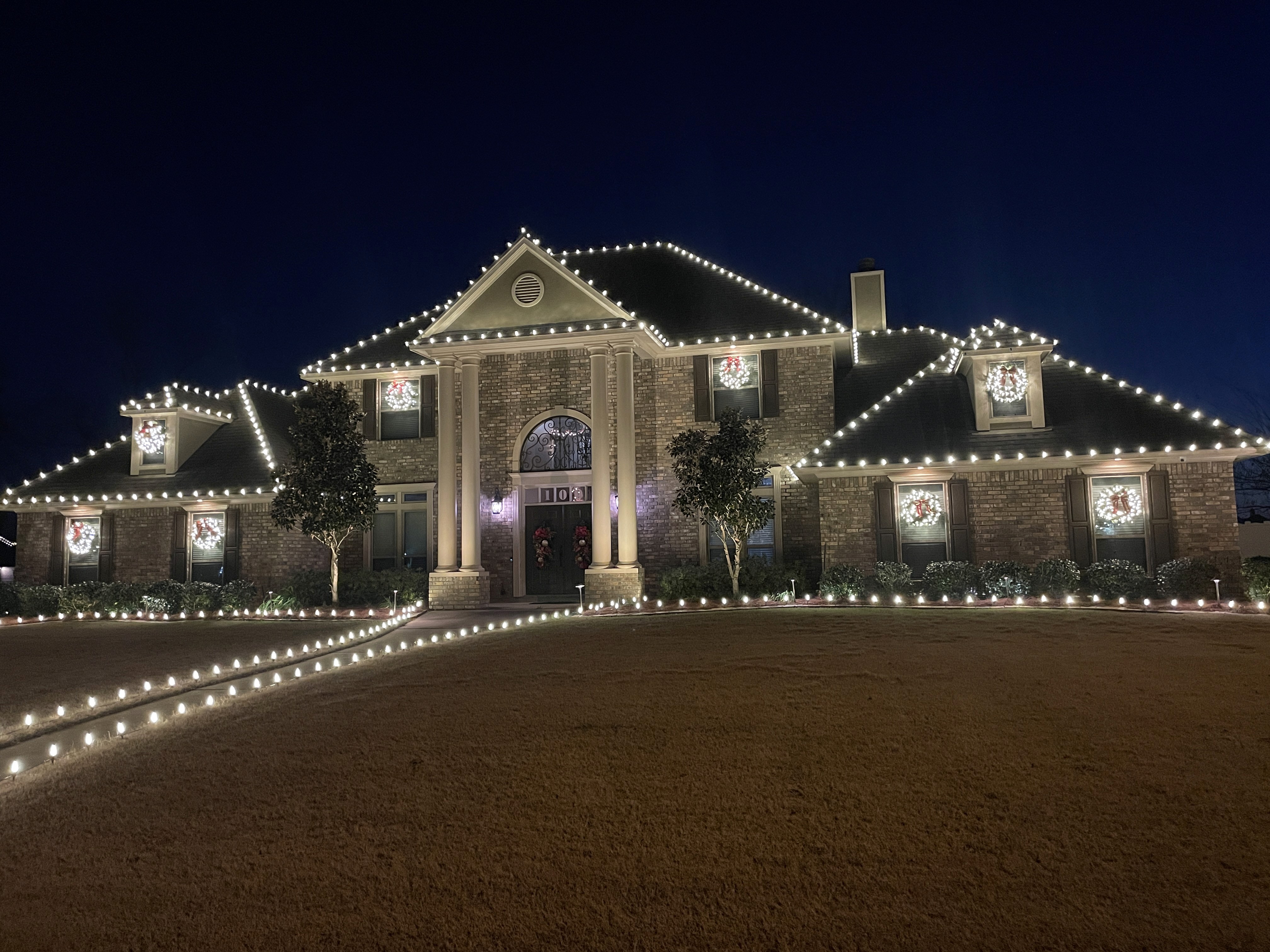Shreveport home with professional Christmas light installation featuring colorful roofline lights, lit bushes, and wrapped tree.