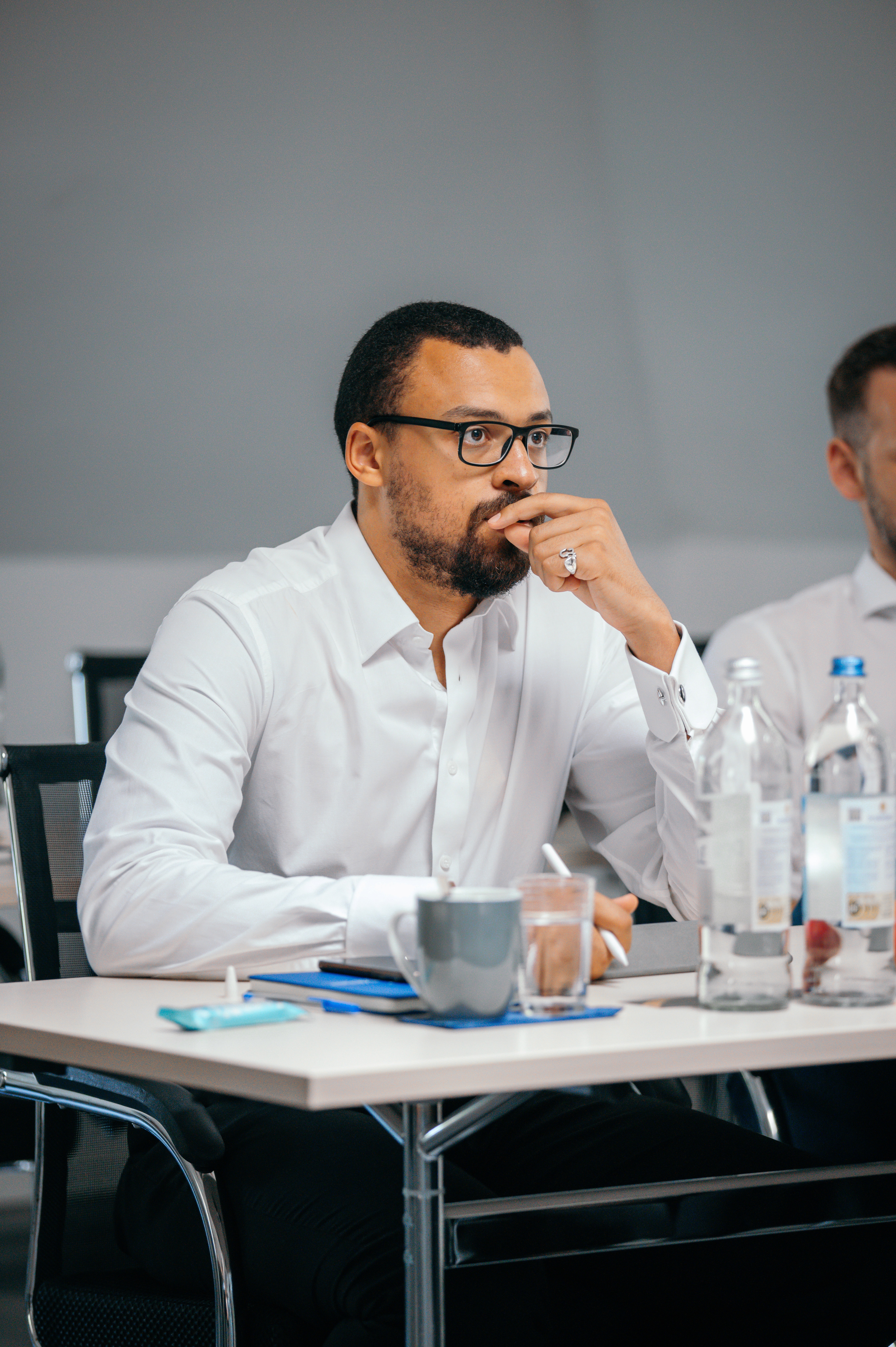A man wearing glasses and a blazer working at a desk, writing in a notebook beside a computer monitor in a modern office.