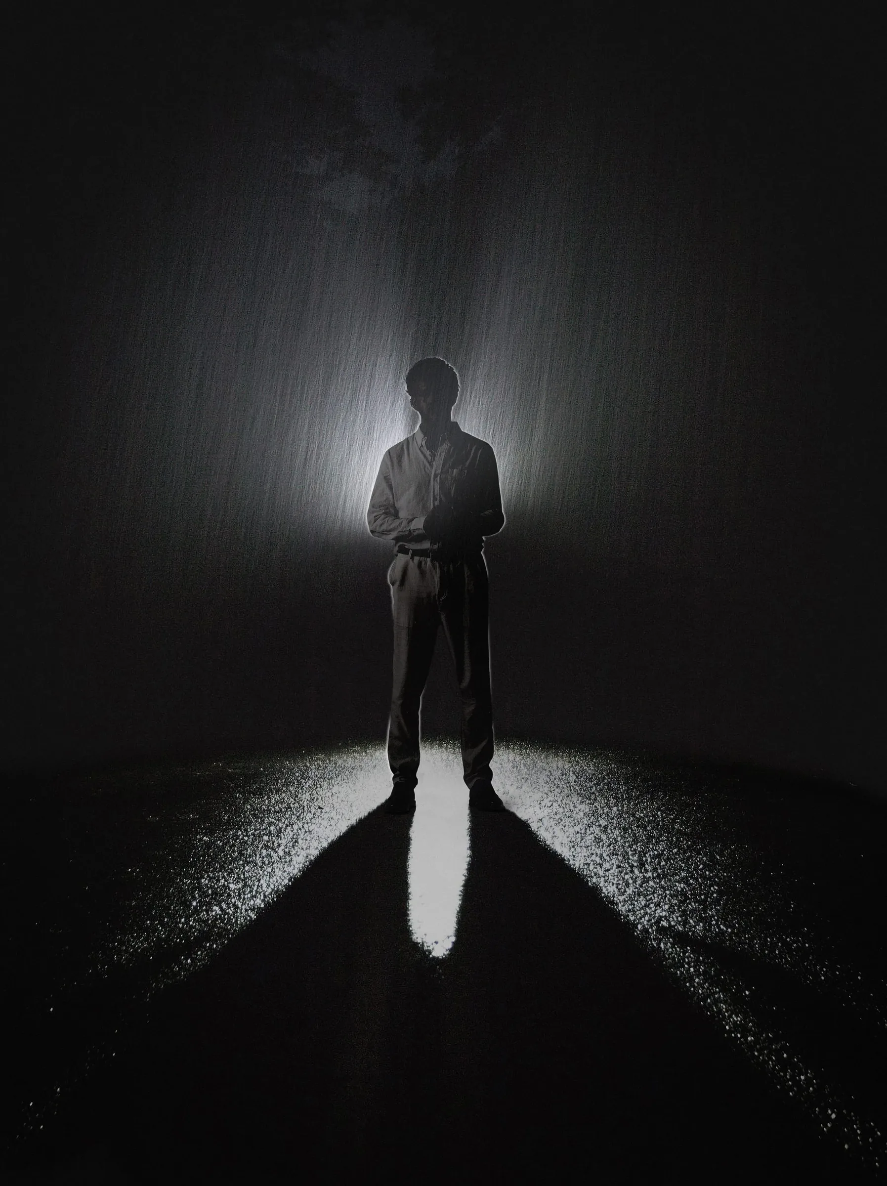 Silhouette portrait of Jeffrey Hakim standing in rain, backlit by a dramatic light beam with a long shadow cast on wet pavement.