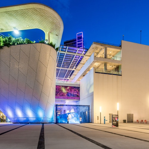 Modernes Gebäude mit geometrischer Architektur, blauer Beleuchtung und beleuchteten Gehwegen unter einem dunkelblauen Abendhimmel.