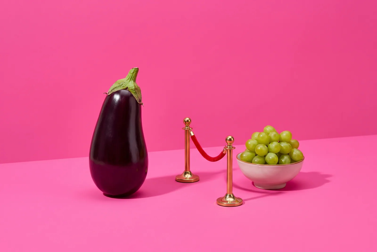 Glossy eggplant standing behind a miniature red velvet rope barrier on a hot pink backdrop, illustrating how to lock YouTube Restricted Mode