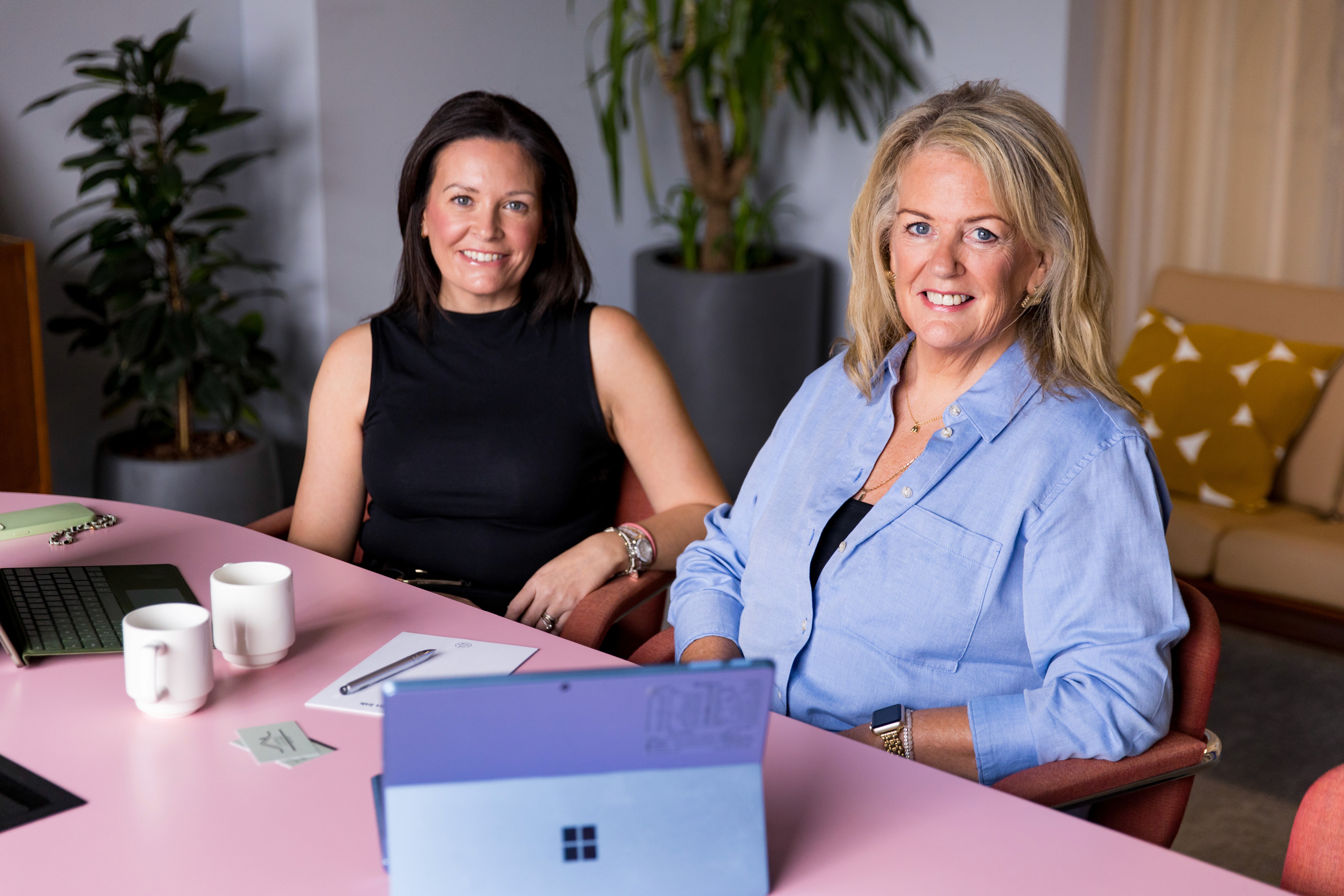 natalie and muriel sat at a pink table together. There is stationary, mugs, and a laptop on the table