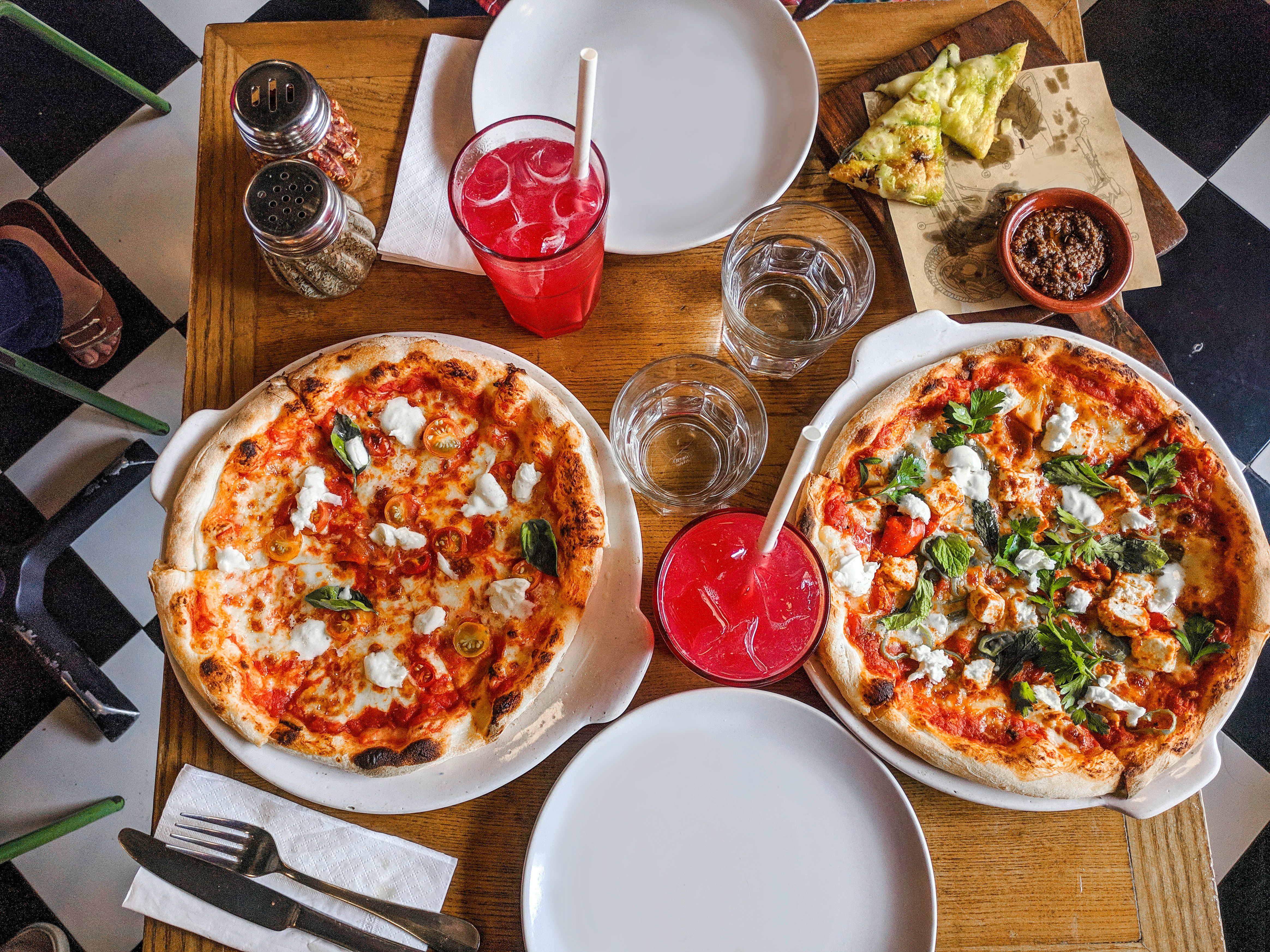Two pizzas on plates with pink drinks on a checkered table
