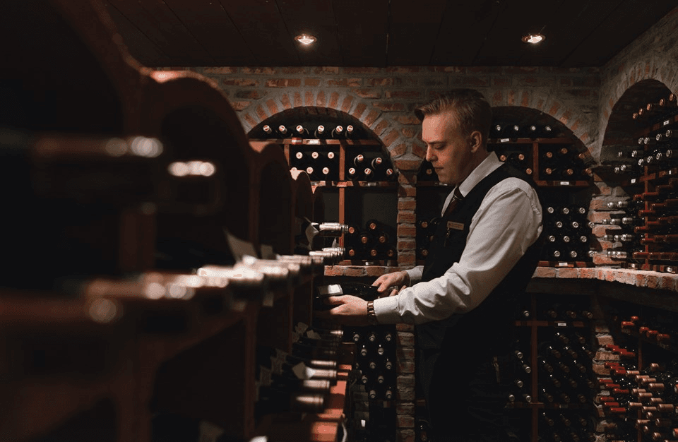 A professional in formal attire is organizing wine bottles in a dimly lit, brick-arched wine cellar, showcasing an extensive collection of diverse wines.