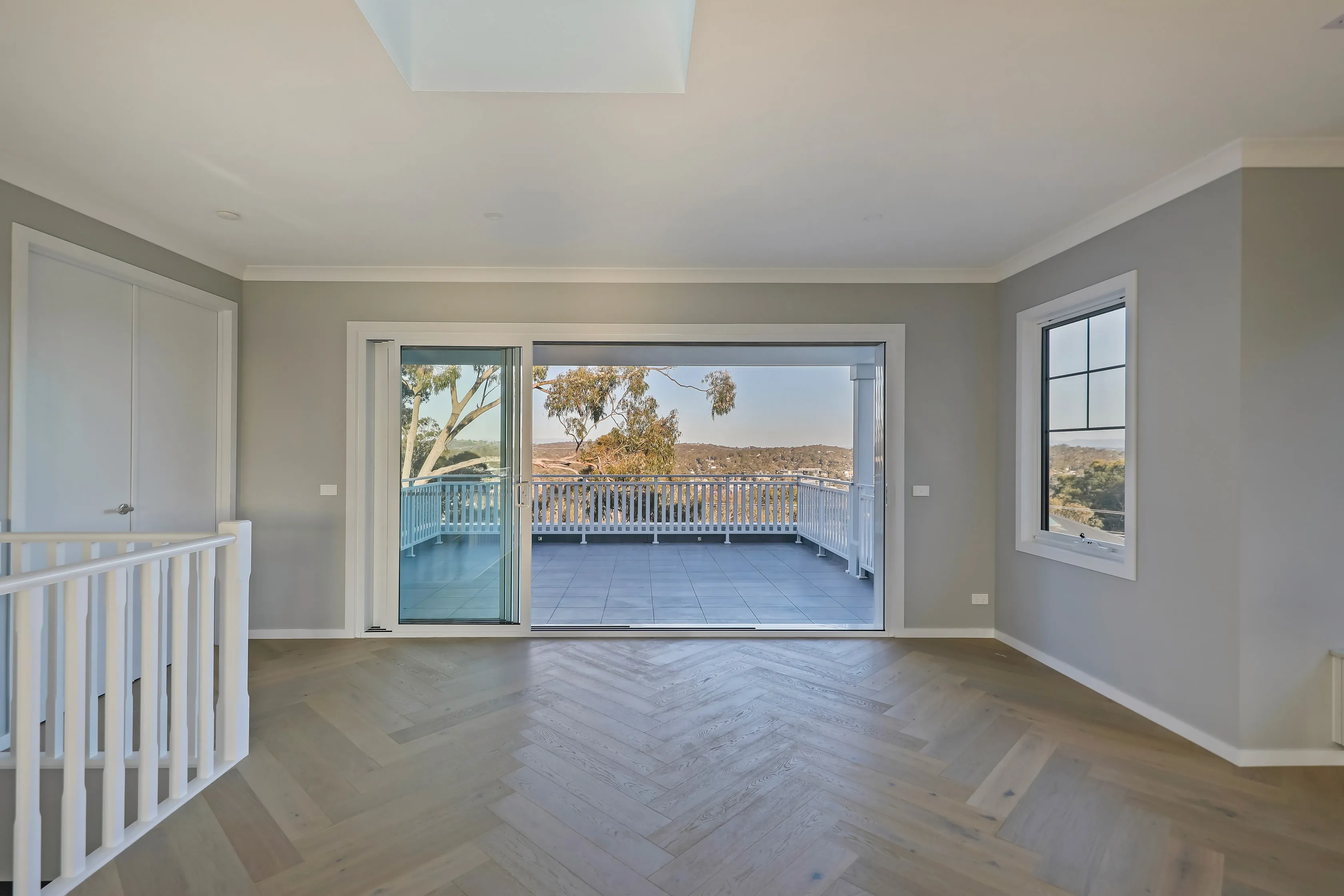 Upstairs retreat with dove-grey walls, oak herringbone floors, a white-balustraded stair void and stacking glass doors opening to a tiled balcony with a treed valley outlook