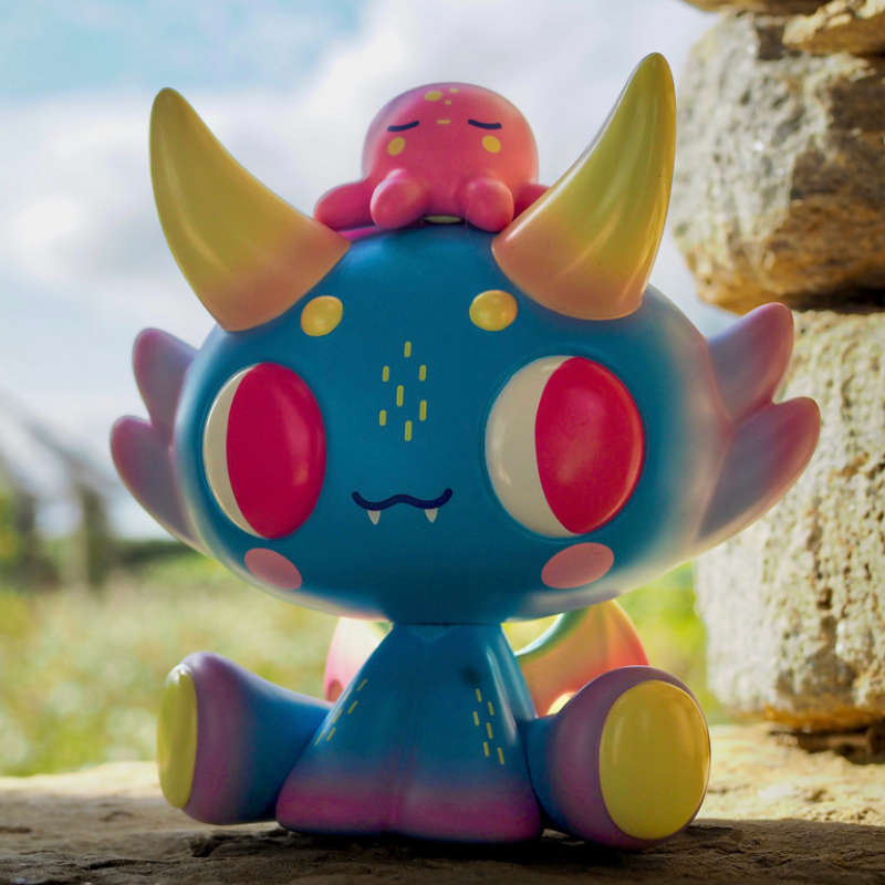 Mostrino by Miguel Guercio. It's a cute blue dinosaur, with a pink octopus on its head, sitting on a stone wall on a bright sunny day.