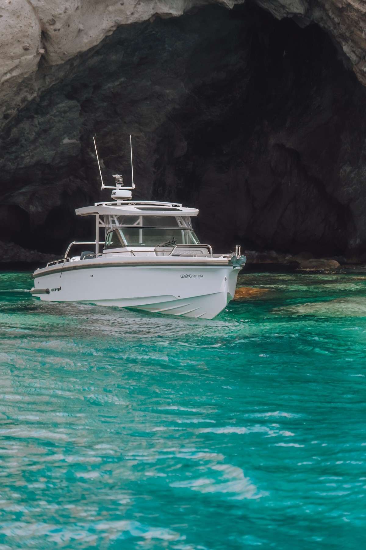 White Axopar 37 motor yacht anchored in crystal-clear turquoise waters beside dramatic limestone cliffs in the Cyclades.