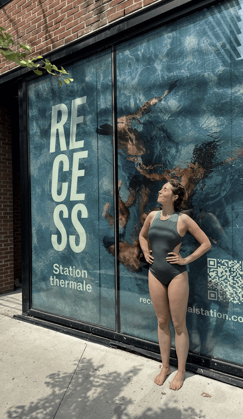 A person stands by the rocky seaside under a clear blue sky, wearing a sleeveless beige t-shirt with the word RECESS printed on the back.
