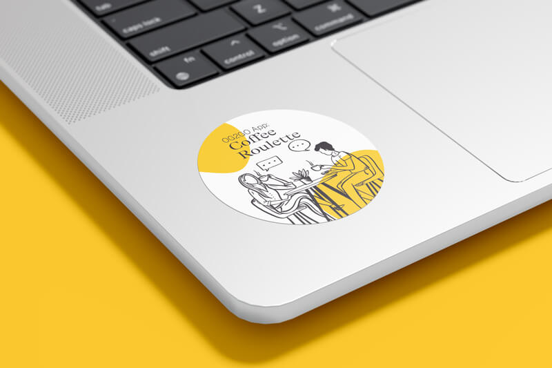 Sticker auf einem Laptop neben dem Mauspad zeigt zwei Menschen an einem Tisch, die sich mit Sprechblasen unterhalten. Darüber steht die Headline „OG2GO App: Coffee Roulette“.