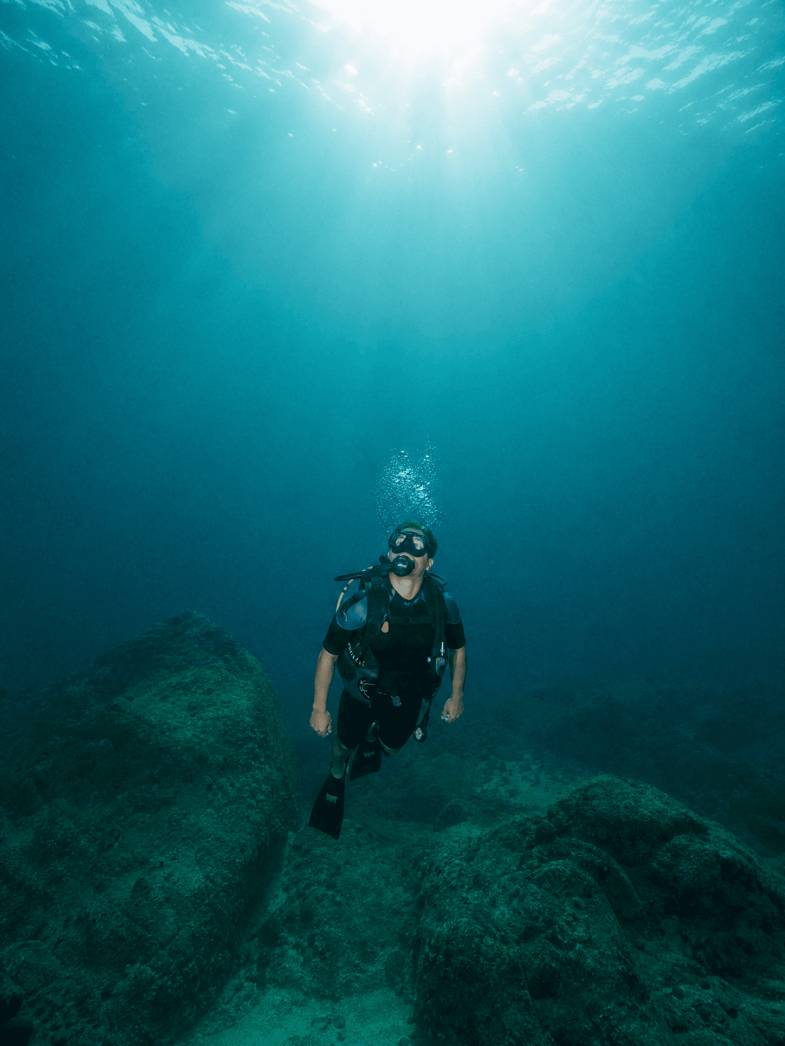 A diver suspended in deep teal water looking up at the light