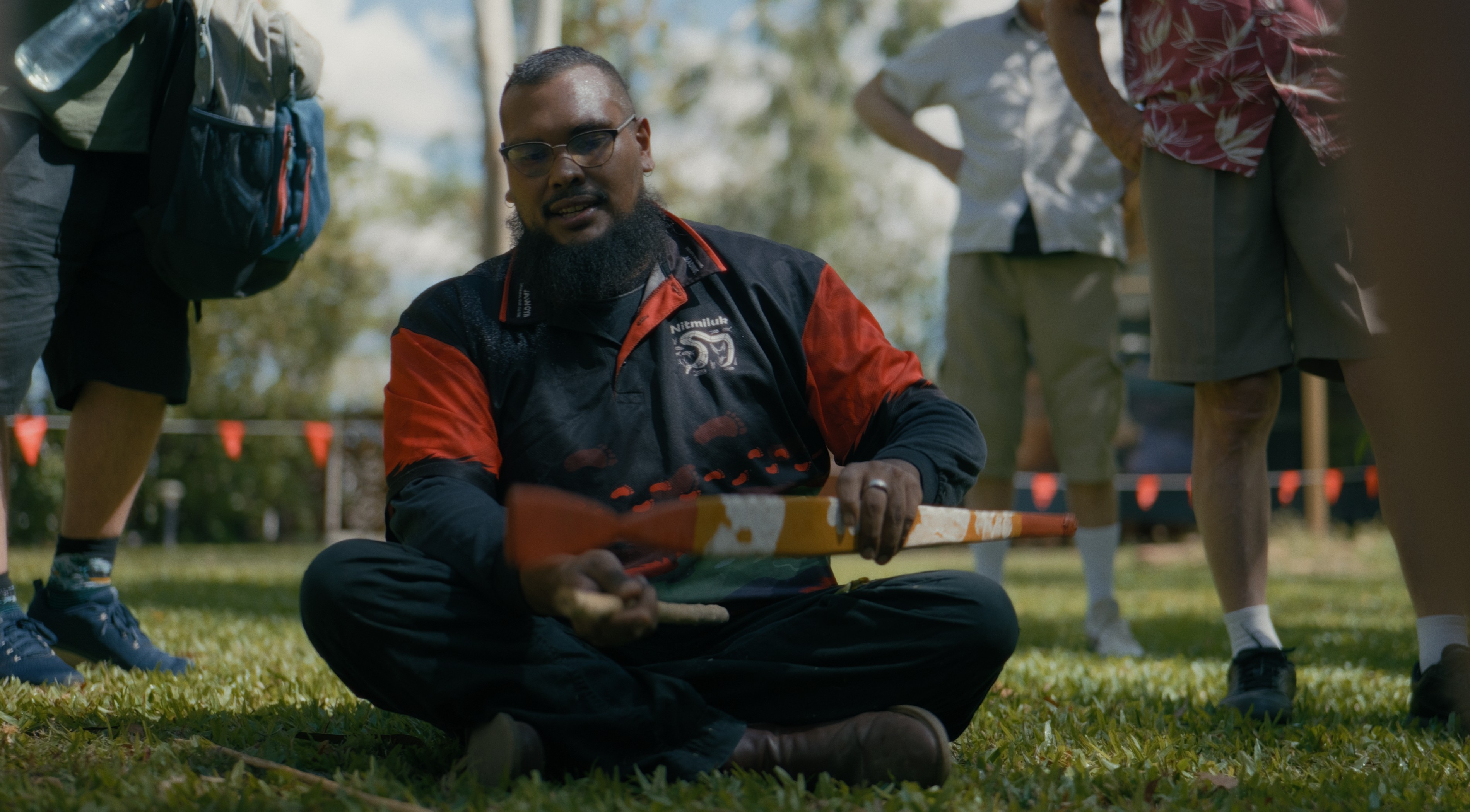 A man sits cross-legged on the grass, holding a boomerang. He's wearing a red and black shirt and speaking to a small group of people.