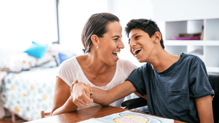 A woman and a young teenage boy share a joyful laugh together at a table, with an open book or drawing in front of them and a bright bedroom visible in the background.