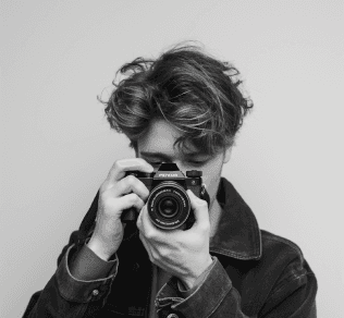 A person with tousled hair is holding a camera up to their face, appearing to take a photo, while wearing a dark jacket against a plain background.