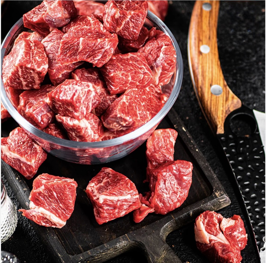 A close-up of a dark gray bowl filled with white fish chunks, cherry tomatoes, green olives, and herbs, served next to a glass of white wine on a white table by a window.