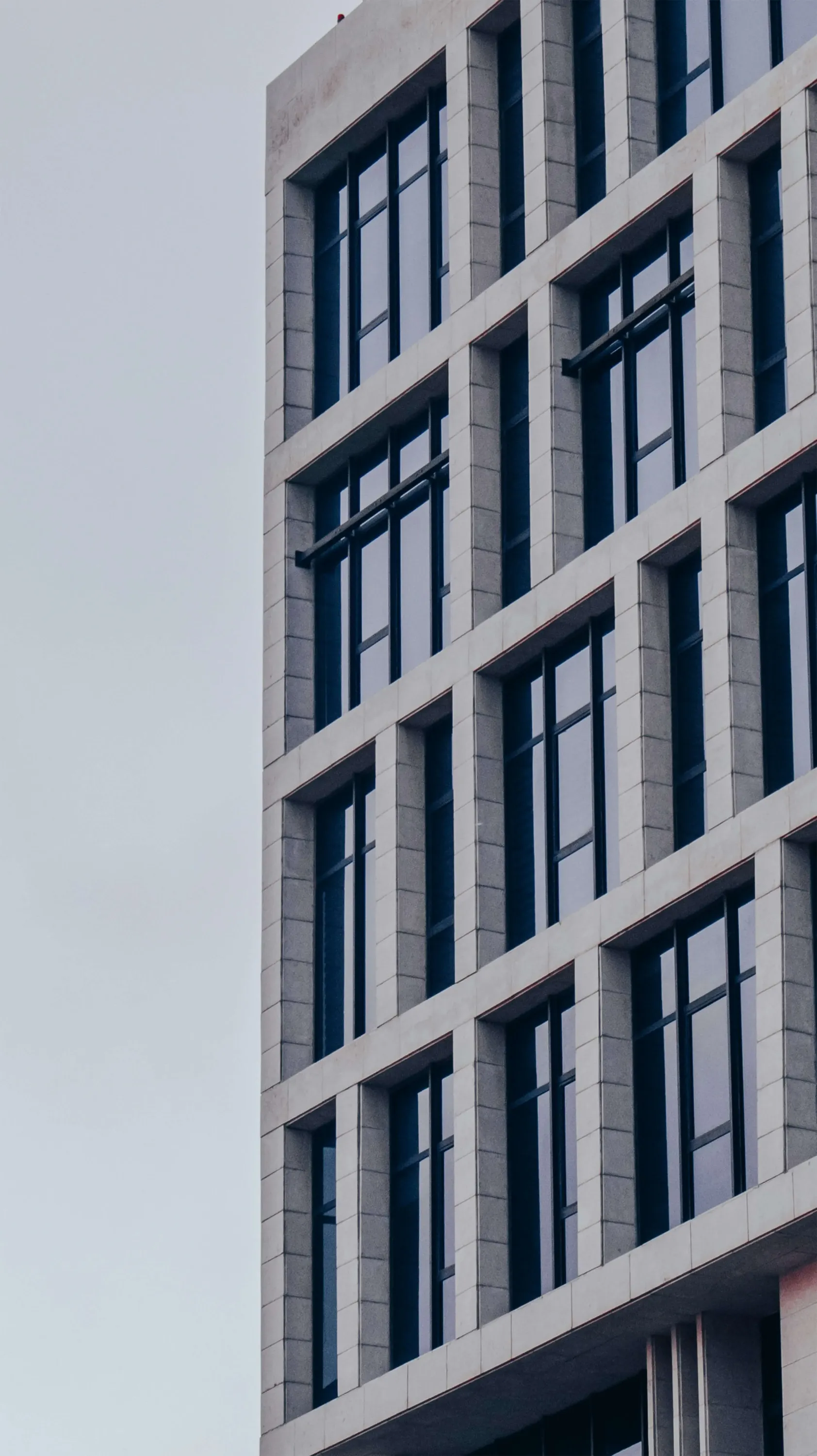 The image shows a section of a modern building facade with a grid-like pattern of large glass windows and prominent stone-colored vertical and horizontal bands against a clear sky.