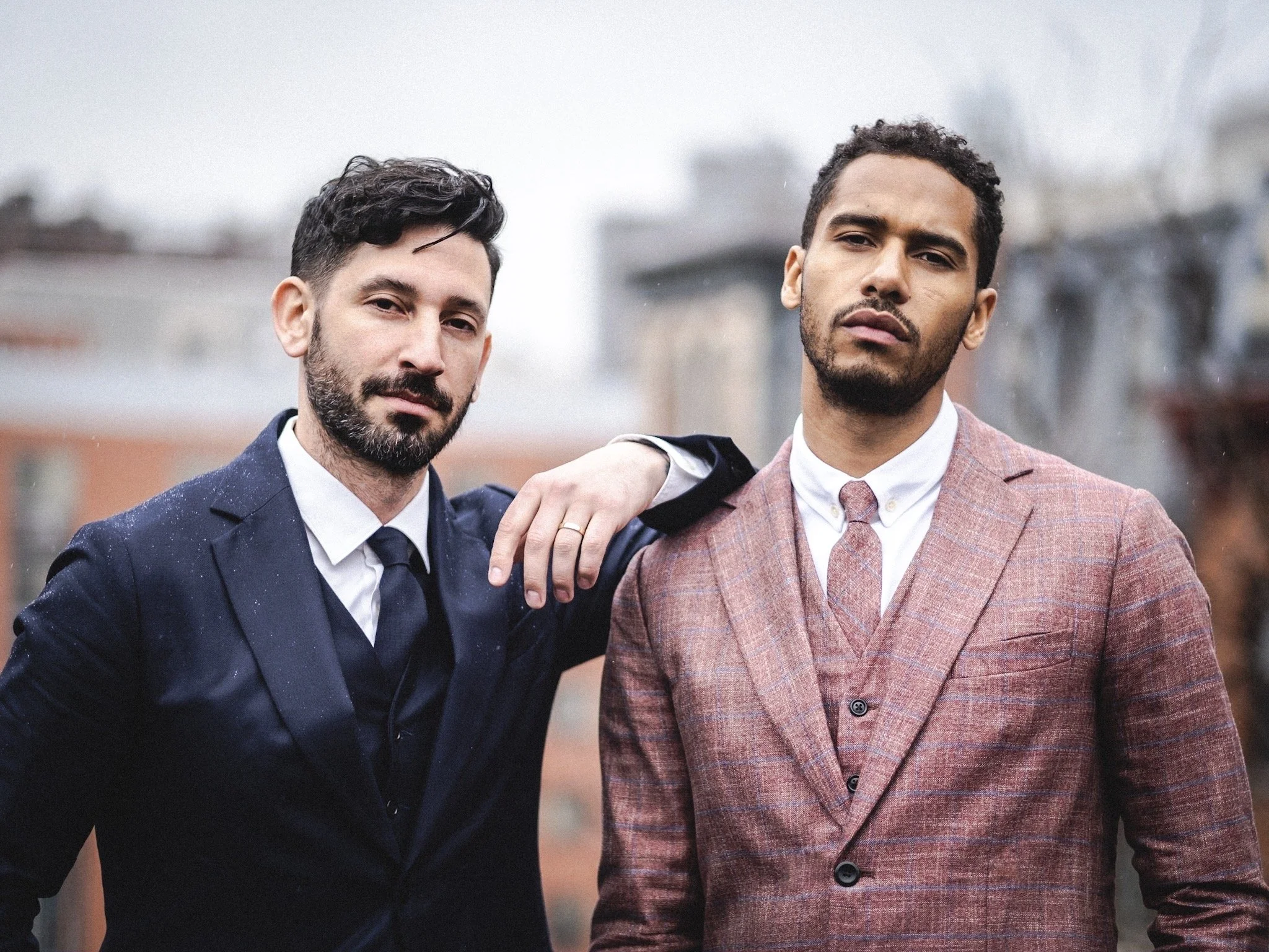 Two men in tailored suits on a city rooftop, one in navy with a beard, one in a rust check three-piece, looking at the camera.