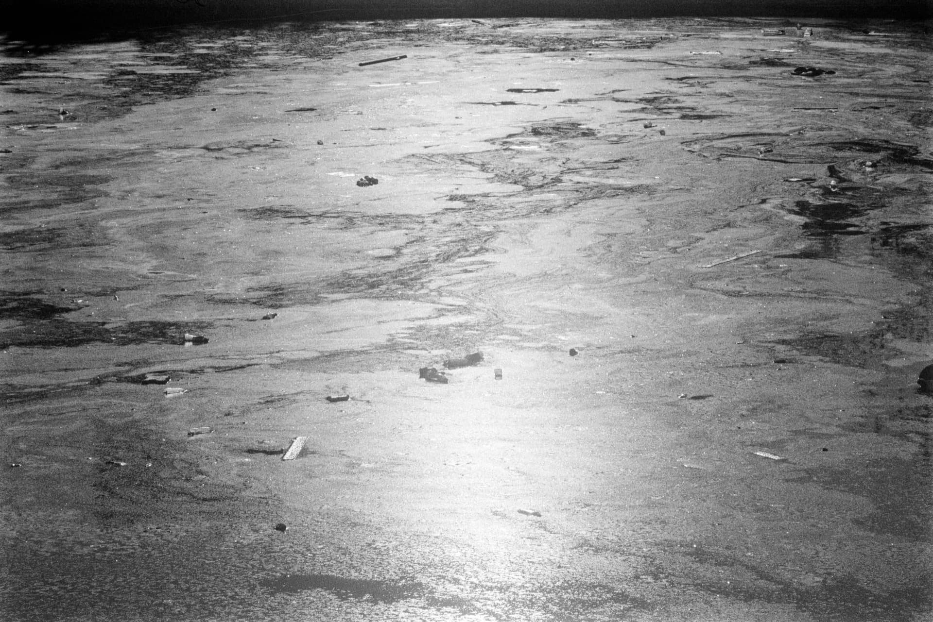 Wide expanse of very still stagnant water, scattered debris floating on reflective surface, horizontal view emphasizing pooling and material decay