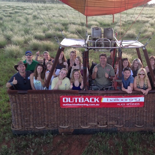 A group of people giving thumbs up in a hot air balloon basket labeled "Outback Ballooning" ready for takeoff over grassy fields.