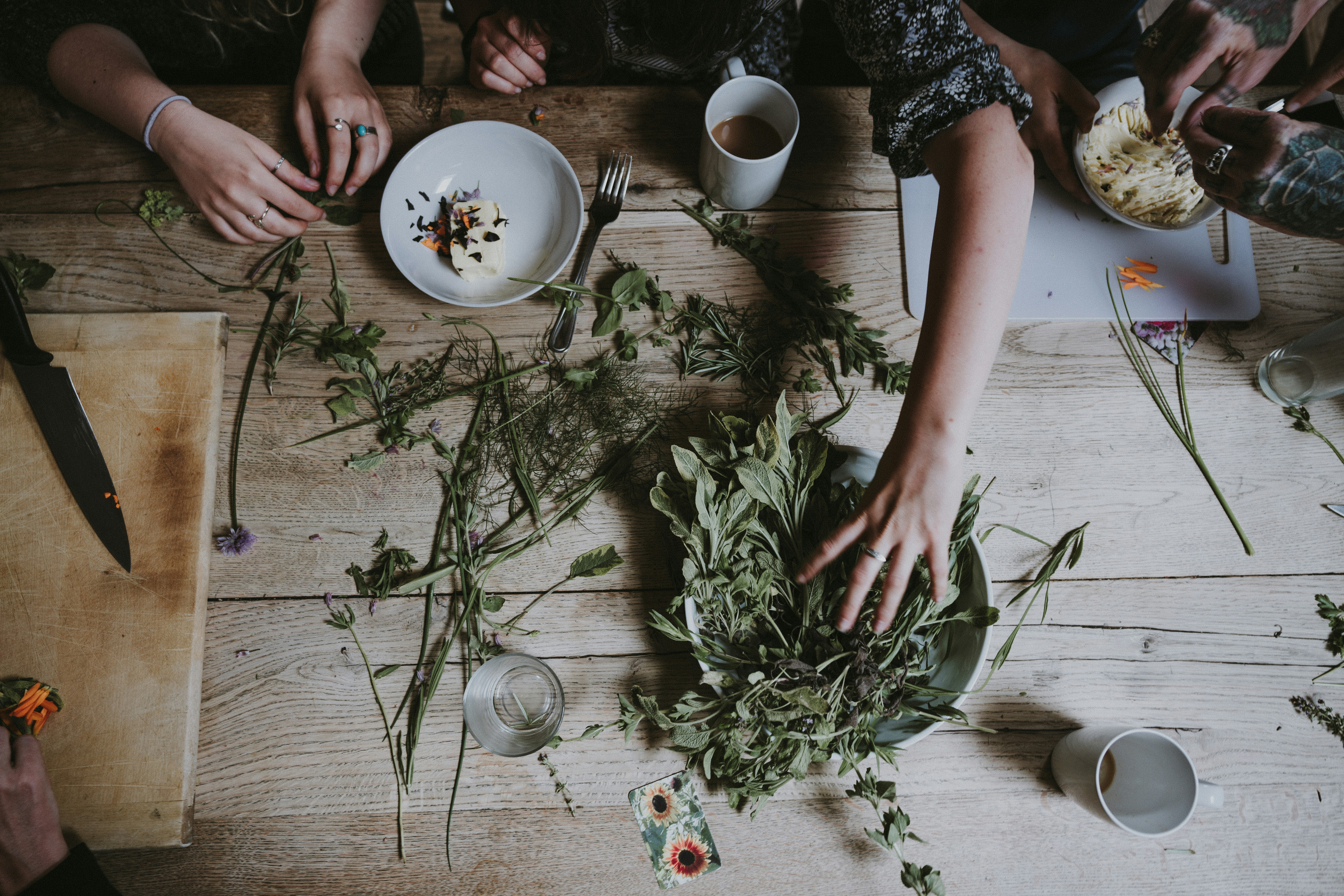 A variety of fresh vegetables and herbs arranged in bowls, including greens, avocados, and spices, on a wooden surface.