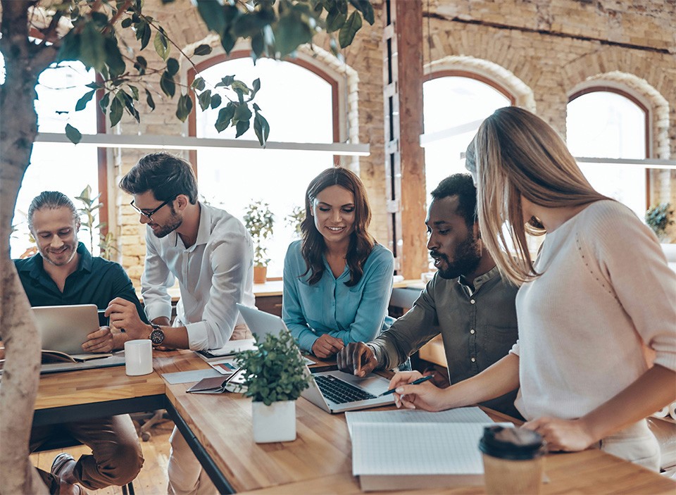 A diverse team of corporate employees in a modern office space, all working together over a table of papers and post it notes