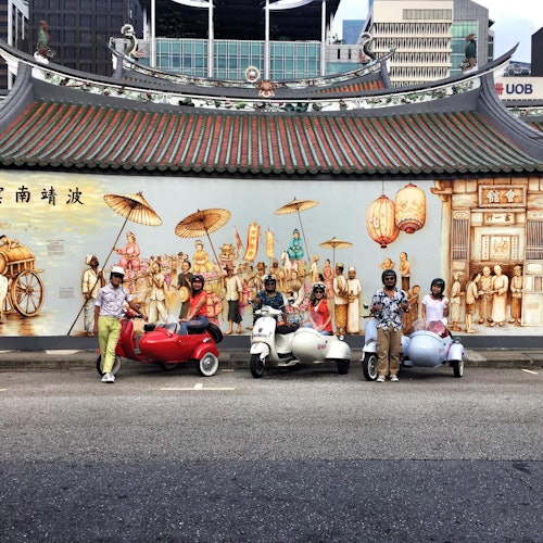 Five people pose beside sidecar motorcycles in front of a mural depicting an old marketplace, with a traditional tiled roof above.