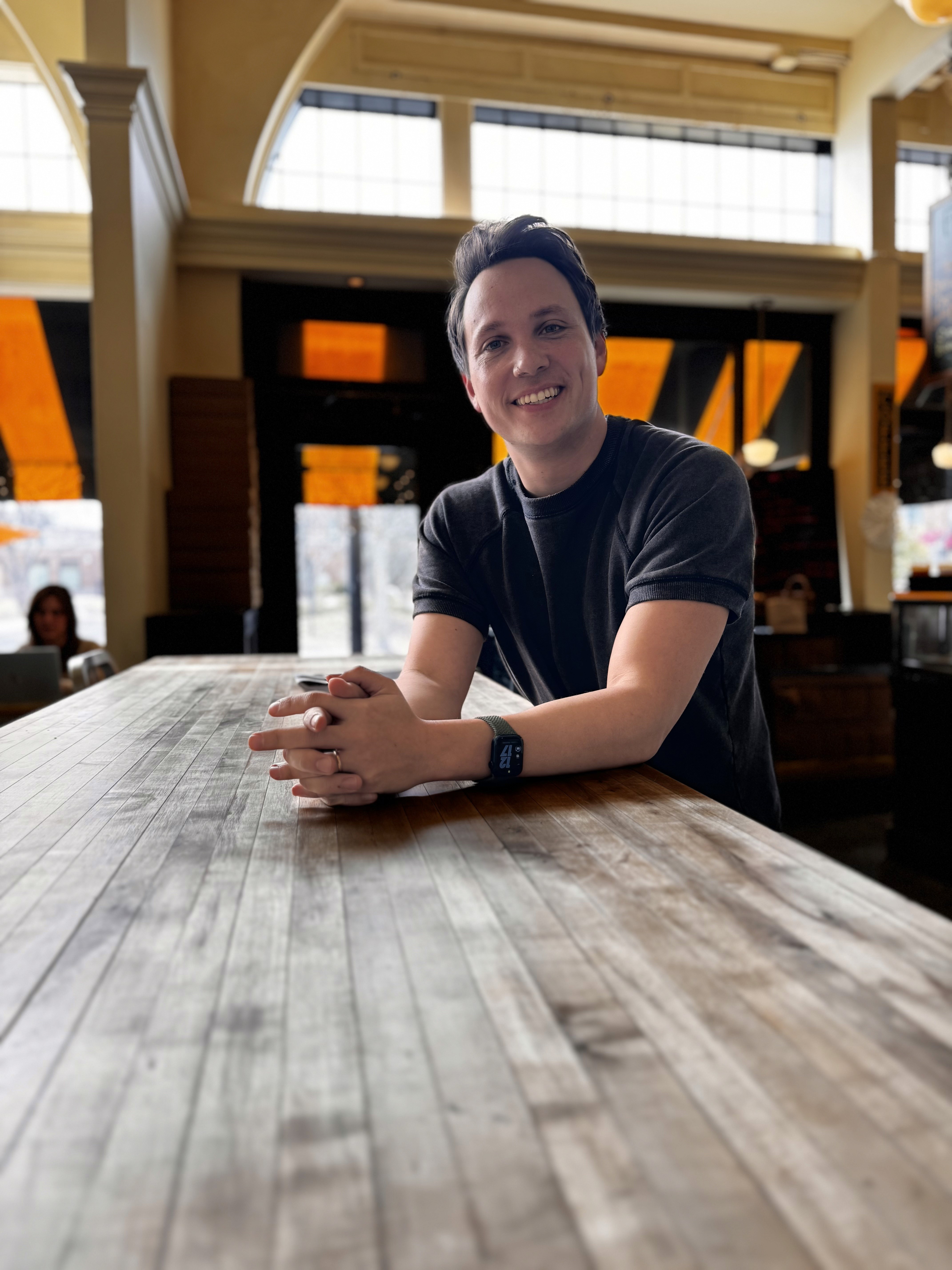 Pastor Zax Calvo smilingl sitting indoors with warm lighting and large windows in the background.