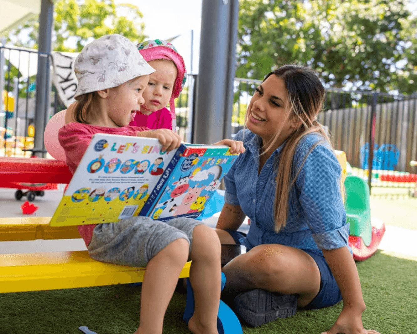 Qualified childcare educator reading with two young children outdoors at Kaleidoscope Kids Early Learning Centre in Brisbane.