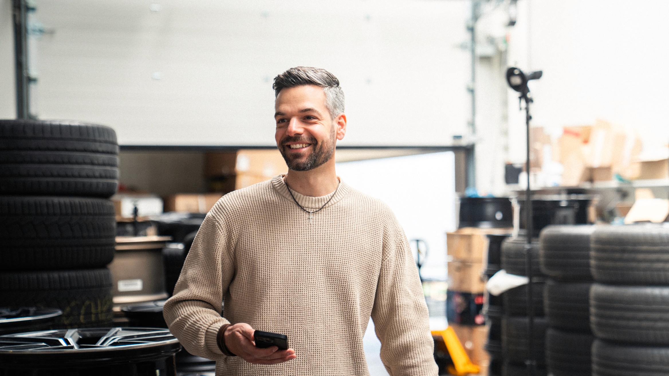 Schwarz-weiß Porträt eines lächelnden Mannes mit Smartphone in der Hand, stehend in einer Werkstattumgebung von Insiderfelgen – symbolisiert Kundennähe und professionelle Beratung.