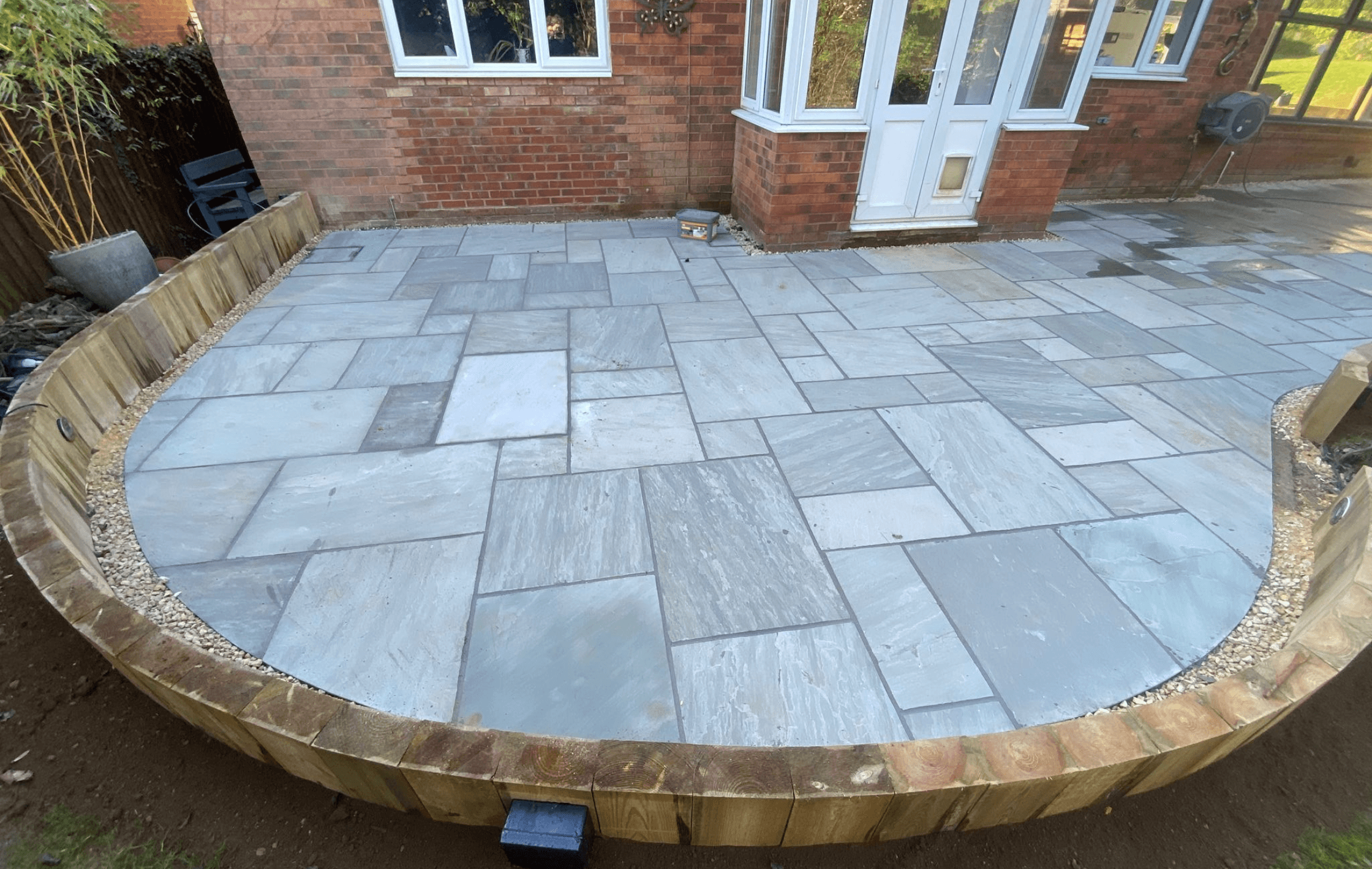 Bespoke Indian sandstone patio installation by PPM Builders in Northampton. Expertly designed outdoor paving with a curved edge and wooden sleeper border, enhancing garden aesthetics and functionality.